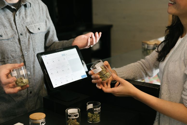 Two people, one a dispensary employee and one a customer, evaluate jars of cannabis flower at the checkout counter. A tablet with checkout options is displayed on the screen.