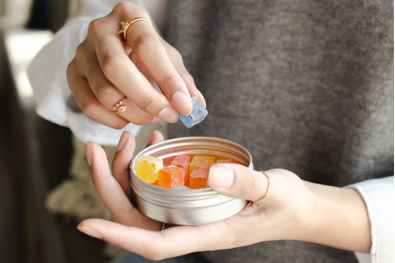 A close up of a pair of hands holding a tin of gummies. One hand is holding a gummy, while the other holds the open tray. This photo illustrates New Jersey's expansion of its edibles program.