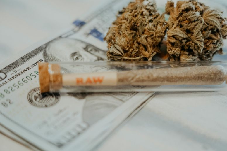 Dried cannabis flower and a pre-roll in a clear case are positioned on top of $100 bills. This illustrates some of the major financial news to come out of New York cannabis in January 2024, including first year sales numbers and proposed tax revisions.
