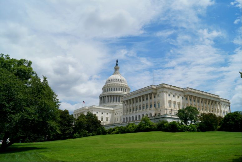 A photo of the Capitol Building. The building is captured at an angle. It's against the backdrop of a blue sky with fluffy white clouds. A green lawn and trees are pictured in the foreground.