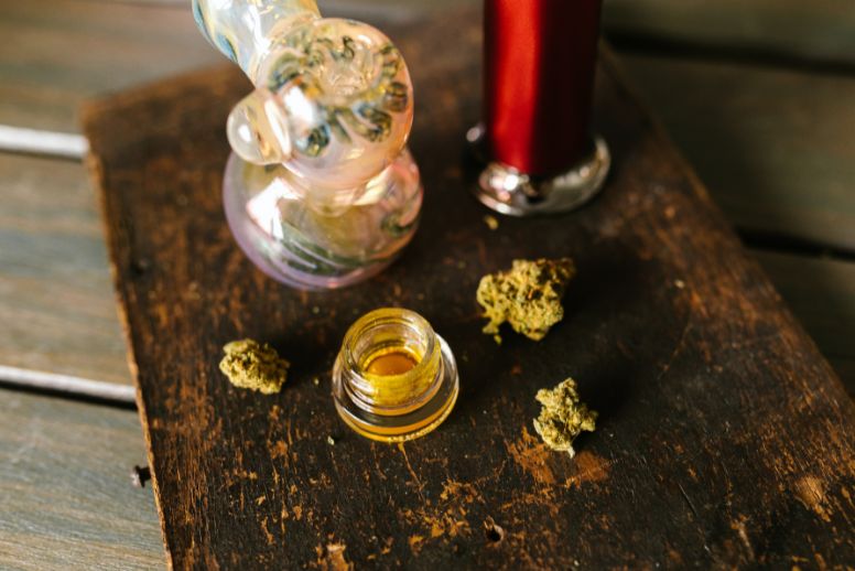An open container of cannabis concentrates sits on a dark wood table. Bits of cannabis flower surround the concentrate. A glass piece is seen in the background.