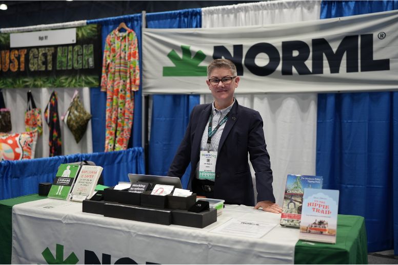A representative from the National Organization to Reform Marijuana Laws (NORML) stands at their booth at CWCBExpo 2023. The representative is standing in the center of the photo, wearing a dark suit jacket and a light shirt. The NORML logo is visible in the background and foreground.