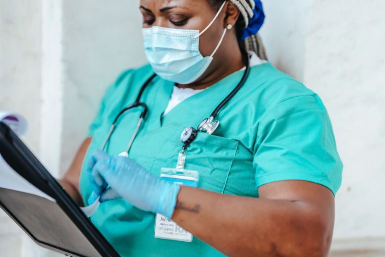 A nurse wearing blue-green scrubs, a mask, and blue gloves examines a patient chart. This photo illustrates the approval of cannabis nursing as a specialty by the American Nursing Association in October 2023.