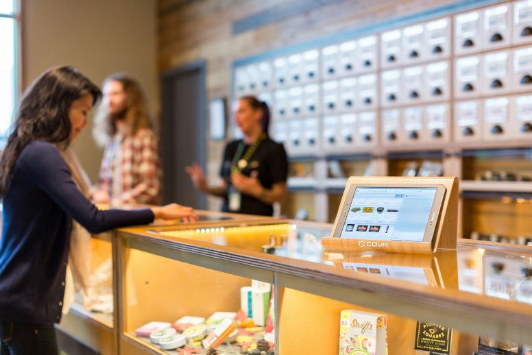 A photo inside a dispensary. A point of sale system on a tablet is on display in the foreground, open atop a glass case. Three people can be seen out of focus shopping and assisting a customer in the background.