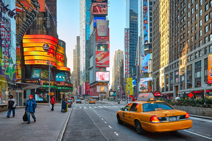 A shot of New York City's Times Square. Multiple billboards and tall buildings can be seen. A yellow taxi is in the foreground.