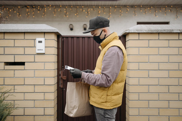 A delivery person wearing a yellow puffer vest and a black baseball cap delivers a bag to a home with a gate.