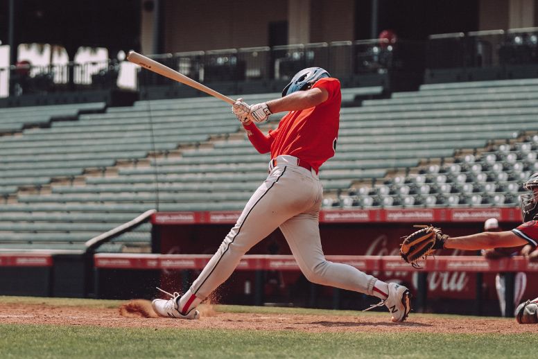 A baseball player mid-swing against the backdrop of a stadium. The player is wearing a red jersey and white pants.